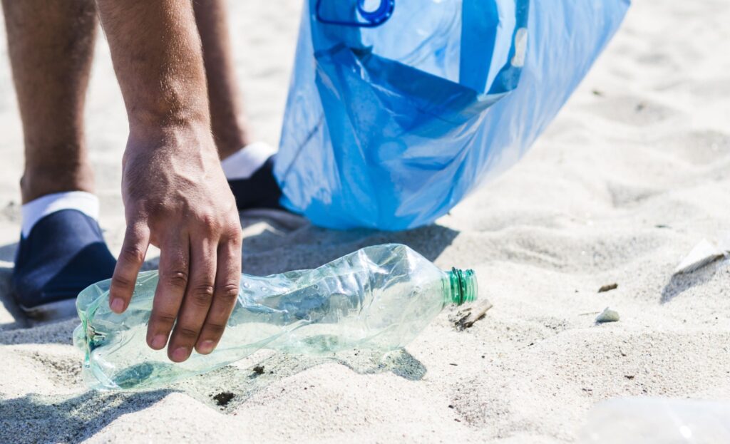 Pessoa recolhendo garrafa plástica da areia durante ação de limpeza em praia