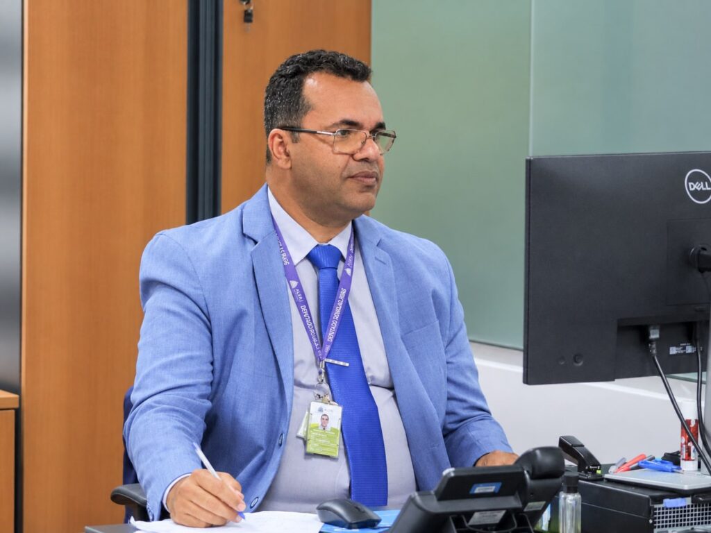 Edinaldo Basílio sentado à mesa de trabalho, com paletó e gravata azuis